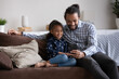 © fizkes - Positive happy young Black dad hugging cute daughter girl with love, tenderness, using cellphone on couch at home, making video call, taking selfie. Father and kid posing on sofa for selfie