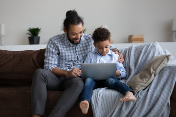 Positive happy young African father hugging zen Z son kid. Excited dad giving support to little gamer child playing virtual video game on computer. Fatherhood, modern technology in family