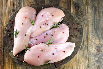 Fresh raw chicken breast fillets sprinkled with peppercorns and rosemary on round wooden board.