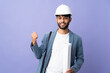 © luismolinero - Young architect Moroccan man with helmet and holding blueprints over isolated background pointing back