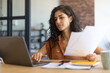 © Prostock-studio - Focused young businesswoman working or studying on laptop, holding documents, sitting at desk indoors, copy space