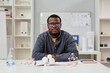 © Seventyfour - Front view portrait of African American teacher sitting at desk in chemistry class with molecule models, copy space