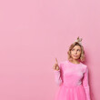 Upset displeased young European woman has hairstyle with disappointed expression wears festive dress and crown points above at copy space shows something unpleasant poses against pink background.