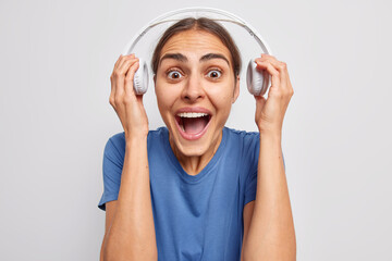 Wall Mural - Emotional dark haired woman takes off headphones listens music with loud sound exclaims and keeps mouth widely opened stares bugged eyes dressed in casual blue t shirt isolated over white background