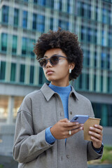 Wall Mural - Attractive young curly haired woman wears sunglasses and stylish jacket uses cellphone chats in social networks drinks coffee poses outdoors near modern building. People lifestyle and technology