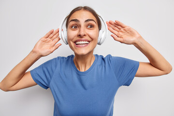 Wall Mural - Positive young European woman with combed dark hair keeps arms raised smiles broadly listens favorite music via wireless headphones dressed in casual blue t shirt isolated over white background.