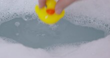 Person Filled A Tub With Hot Water And With Soap Bubbles, Preparing It To Take A Relaxing Bath Or To Bathe A Child, So He Threw Yellow Toy Rubber Duck Into The Water, Close Up