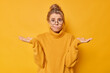 Indoor shot of hesitant clueless young woman spreads palms cannot make decision shrugs shoulders dressed in casual jumper isolated over yellow background. Questioned female model feels uncertain
