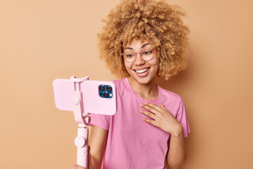 Wall Mural - Glad positive woman with blonde curly hair shoots video via smartphone on selfie stick records blog wears casual pink t shirt isolated over beige background. People and modern technologies concept