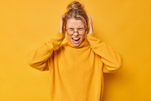 Waist Up Shot Of Emotional Young Blonde Woman Screams Loudly Covers Ears With Hands Doesnt Want To Hear Bothering Sound Wears Spectacles And Sweater Isolated Over Yellow Background. Stop This