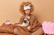 © WHstudio Leushin N - Displeased young woman with curly hair wrapped in blanket dressed in slumber suit and blindfold on forehead poses with pug dog in bed has bad mood awakes early isolated over brown background.
