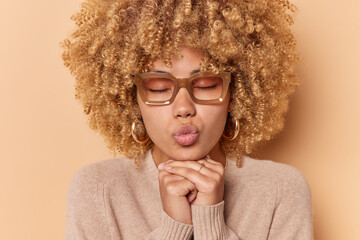 Wall Mural - Headshot of beautiful young woman with curly bushy hair keeps hands under chin lips folded has romantic facial expression wears earrings nd casual jumper isolated over beige background sends mwah