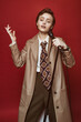 Fashionable beautiful singer girl in retro coat, tie and pants with microphone in hands posing on red background. Vintage toning.
