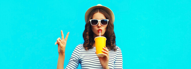 Wall Mural - Portrait of stylish young woman drinking a fresh juice wearing a summer straw hat and striped t-shirt on blue background