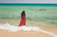 Woman In Red Dress Walking On Tropical Beach Towards Shore And Turquoise Blue Ocean In Sardinia