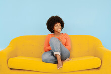 Happy Woman Sitting On Yellow Sofa In Front Of Blue Background