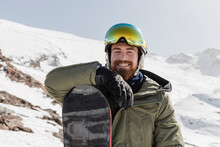 Smiling Young Man Wearing Ski Goggles Standing With Snowboard On Sunny Day