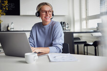 Happy Blond Businesswoman Wearing Headset Sitting With Laptop At Table