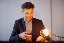 Businessman Controlling Light Bulb With Smart Phone At Work Place
