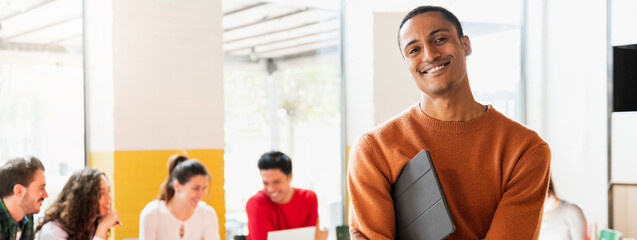 Horizontal banner or header smiling African handsome young man holding tablet and looking at camera. Group of people sitting ad desk on background.