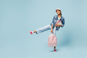 Wall Mural - Full body fun little schoolgirl kid teen girl of African American ethnicity 12-13 year old in denim jacket backpack cap hold exercise books raise up leg isolated on pastel plain light blue background.