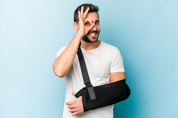 Wall Mural - Young caucasian man with broken hand isolated on blue background excited keeping ok gesture on eye.