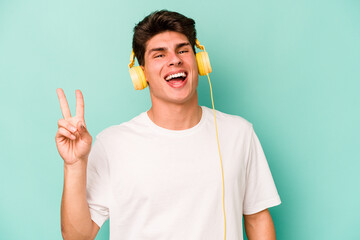 Young caucasian man listening to music isolated on blue background joyful and carefree showing a peace symbol with fingers.