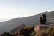 © KAMPUS - Rear view of young backpacker sitting on stone looking at view. Woman with backpack resting on top of mountain. Hiking concept