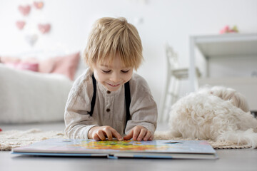 Wall Mural - Cute blond preschool child, blond boy with pet maltese dog, reading book at home with mother