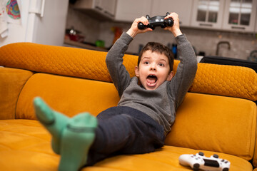 Cute 5 year old boy playing video game console while sitting on orange couch at home.