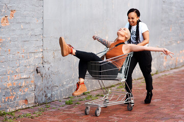 Canvas Print - Im about to fly. Full length shot of an energetic young woman pushing her female friend in a shopping cart outdoors.