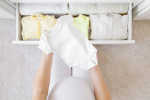 Young Adult Pregnant Woman With Big Belly Sitting On Carpet. Hands Holding Bodysuit And Sorting Clothes For Future Baby In White Drawer Box. Preparing Things In Pregnancy Time. Top Down View. Closeup.