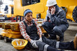 © pressmaster - Young female engineer with hurting knee sitting on the floor by anxious male worker in safety helmet and uniform calling ambulance