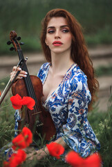 Wall Mural - Beautiful romantic girl with red hair and blue dress holding violin on nature field of flowers. Photo of sensual woman.
