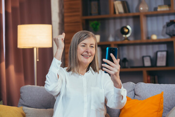 Happy senior gray-haired woman got good news online, sitting on sofa at home and rejoices celebrating victory looking at phone screen