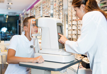 Wall Mural - Positive friendly smiling teenage boy receiving eye examination on modern refractometer in optical store with professional diligent serious female ophthalmologist