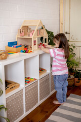 Montessori material. Child girl in pink T-shirt arranges wooden furniture in a doll house.