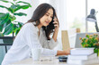 © Bangkok Click Studio - Asian young beautiful professional successful smart female businesswoman on call via smartphone while working with data information on laptop computer writing note on notebook at workstation desk