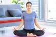 Asian young happy peaceful calm female model in casual sporty outfit sitting crossed legs in lotus position on yoga mat learning studying online meditation class via laptop computer in living room