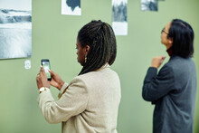 Young African American Woman Attending Exhibition In Modern Art Gallery Scanning QR Code Using Smartphone To Find More Information About Photo On Wall