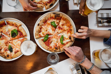 Wall Mural - Top view restaurant table with Italian Margherita pizza and glasses of beer. Unrecognizable multiracial people's hands grabbing a sliced piece.