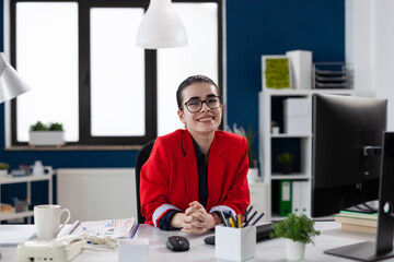 Wall Mural - Portrait of businesswoman with glasses in startup office. Professional entrepreneur in red jacket looking confident in business workspace. Confident employee posing in front of desktop computer.