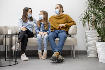 Young family with ten year old girl sitting on a couch at the waiting room of the clinic. Patients in face mask waiting for a doctor's appointment at reception