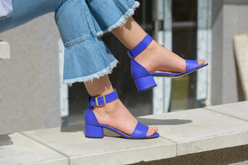 Women's legs in blue denim jeans and sandals in the city street. Trendy elegant casual outfit. Details of everyday summer look.