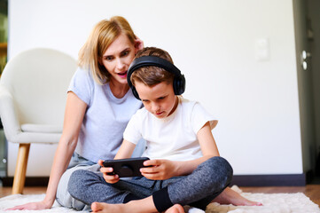 Caucasian boy with mother plays phone on the floor with headphones watching cartoons, listening to music, playing games