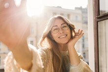 Pretty Caucasian Adult Woman In Glasses Smiling While Looking At Camera While Standing By Open Window On Sunny Day. Blonde In Warm Sweater Spends Time Alone. Beauty, Emotions People Concept