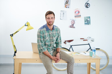 Sticker - Portrait of a casually-dressed young man sitting on his desk in an office. The commercial designs displayed in this image represent a simulation of a real product and have been changed or altered