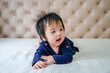 © suppa - Cute little baby girl, playing at home in bed. Cute little caucasian baby lying on bed at home. Little cute baby girl, child in blue dress, smiling happily at camera in white sunny, bright bedroom.