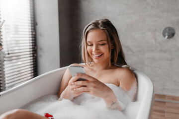 Wall Mural - Happy young lady relaxing in foamy bath, reading beauty blog or watching video online, using smartphone and smiling