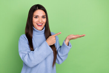 Poster - Photo of pretty excited young woman wear blue sweater smiling pointing finger holding arm empty space isolated green color background